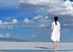 Woman stands alone on a vast salt flat under a bright blue sky with clouds.